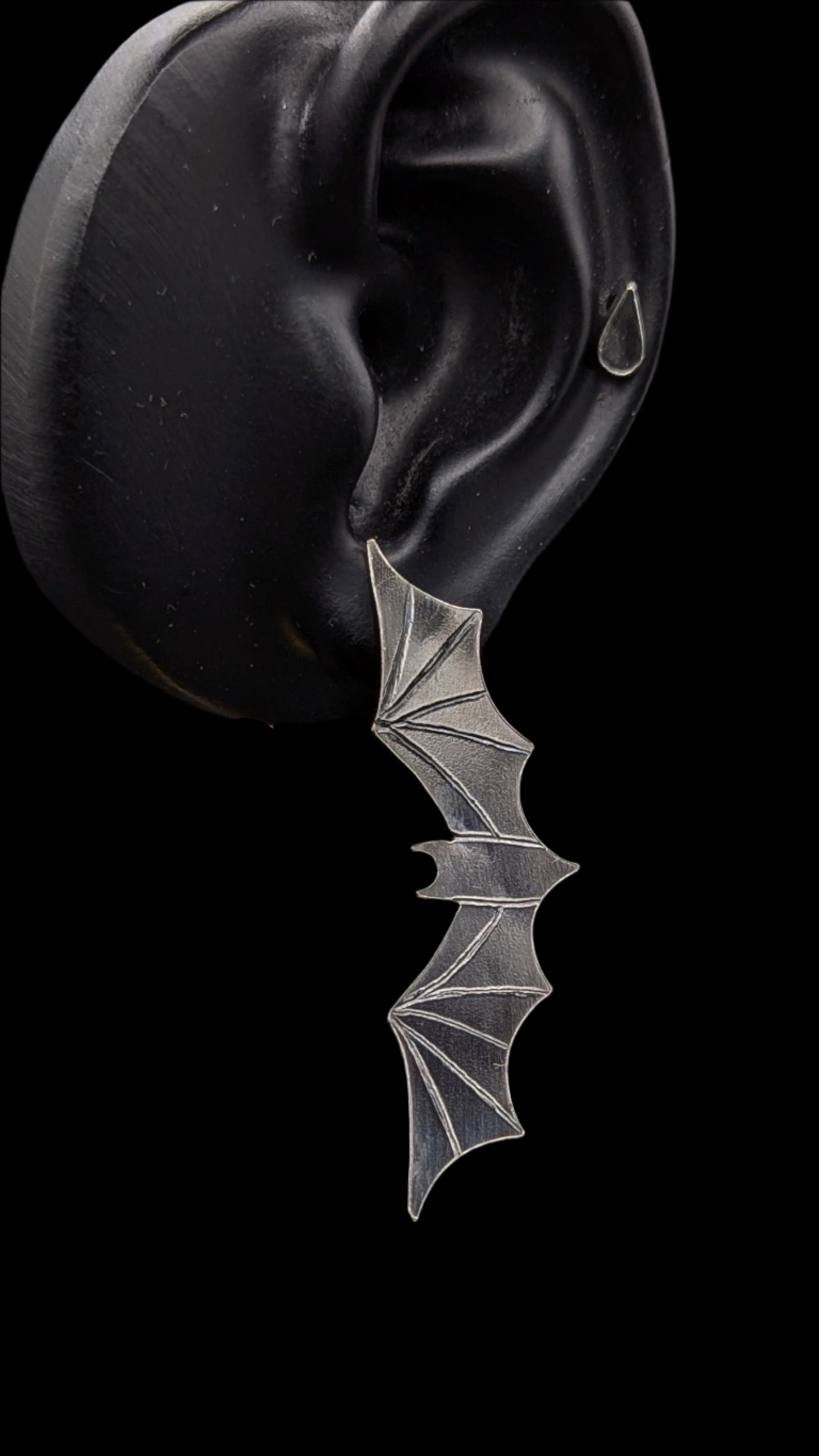Silver bat-shaped earring worn on a black ear against a dark background