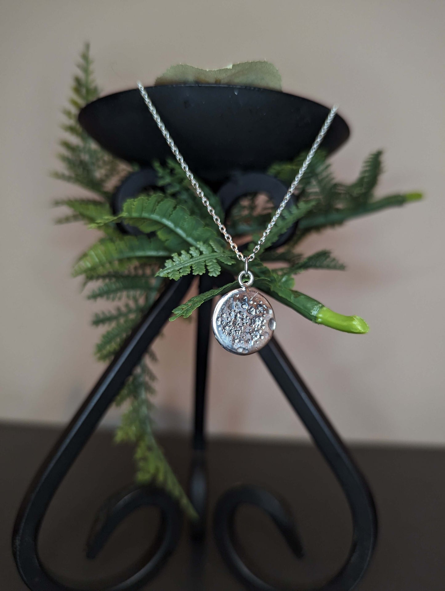 Triluna necklace in the shape of a full moon, hanging from a candle holder together with some leaves